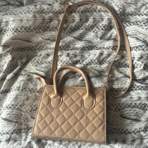 Beige quiltad handväska med axelrem - Snygg beige handväska med quiltat mönster och både handtag och justerbar axelrem. Väskan har en stilren, fyrkantig form och är tillverkad i skinnimitation. Perfekt för dig som gillar klassisk och trendig design.