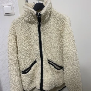 Vit teddyjacka med dragkedja - Mysig vit teddyjacka med hög krage och svarta detaljer. Jackan har två fickor med dragkedja och hel dragkedja framtill. Perfekt för kalla dagar när du vill vara både varm och snygg. Den är använd få gånger men säljer den eftersom jag använder inte den, den ligger bara i garderoben. Storlek M (oversize)