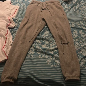 Grå Essentials mjukisbyxor - Snygga grå mjukisbyxor från Essentials Fear of God med elastisk midja och långa snören. Byxorna har tryckt logga på benet och muddar vid bensluten för en skön passform. Perfekta för chill dagar eller streetwear-stil. Dom är helt nya har använt dom cirka 2 till 3 gånger 