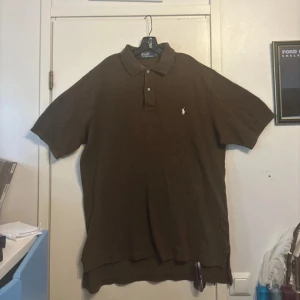 Brun Polo t-shirt från Ralph Lauren - Säljer en brun Polo t-shirt från Ralph Lauren i storlek XLT Tall. Klassisk pikémodell med krage, två knappar och den ikoniska broderade loggan på bröstet. Tillverkad i 100% bomull för skön känsla och snygg passform.