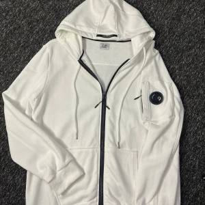Vit hoodie från C.P. Company - Vit hoodie från C.P. Company med svart dragkedja och klassisk linsdetalj på ärmfickan. Tröjan har huva med snörning och ribbade muddar. Perfekt för dig som gillar streetwear och vill sticka ut med en ikonisk detalj.