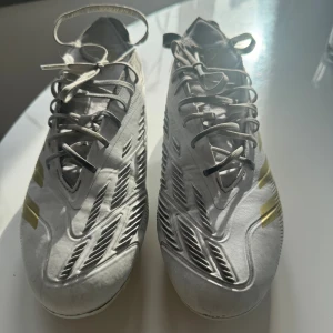 Adidas Predator Elite fotbollsskor vit/guld - Säljer ett par Adidas Predator Elite fotbollsskor i vitt med guldiga detaljer och snörning. Skorna har en modern design med mönstrad ovandel och fasta dobbar för bra grepp på planen. Tillverkade i syntetmaterial och textil för lätt känsla och bra bollkontroll. 