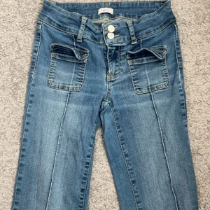 Blå jeans med fickor från Nelly - Snygga blå jeans från Nelly med raka ben och två stora fickor framtill med lock. Jeansen har dubbla silverknappar i midjan och klassiska bakfickor. Materialet är jeans med ljus tvätt, strechiga och markerade sömmar för en fin look.