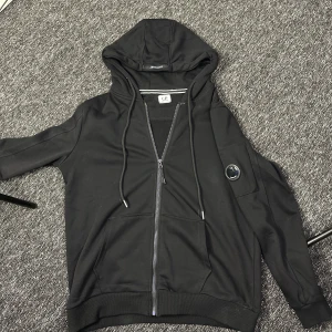Svart hoodie från C.P. Company - Svart hoodie från C.P. Company med dragkedja, huva med snörning och klassisk linsdetalj på ärmen. Tröjan har känguruficka framtill och är tillverkad i mjuk bomullsmix. Perfekt för en chill och stilren look.