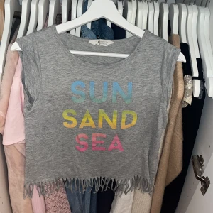 Grå croppad linnetopp H&M med fransar❤️ - Grå croppad linnetopp från H&M med färgglad text 'SUN SAND SEA' i blått, gult och rosa framtill. Skriv för fler bilder❤️🫰🏼🫰🏼