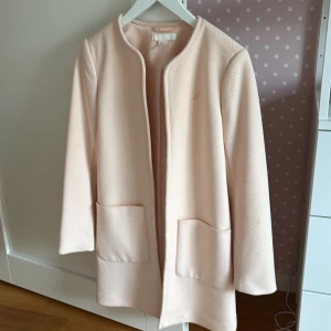 Ljusrosa kappa med fickor från H&M - Snygg ljusrosa kappa med rak passform och två stora fickor framtill. Jackan har rundad halsringning och är helt utan knappar eller dragkedja. Kappan är i storlek 34.