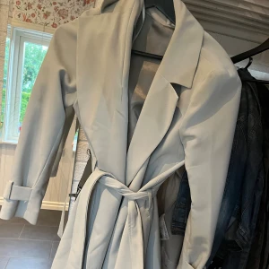 Ljusblå jacka med bälte, strl S - Snygg ljusblå trenchcoat i klassisk modell med bred krage, knytskärp i midjan och dekorativa detaljer vid ärmsluten. Jackan har långa ärmar och en stilren look som passar perfekt till vår och höst. 