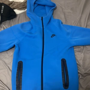 Blå Nike hoodiejacka med dragkedja - Nike hoodiejacka i klarblå färg med hel dragkedja framtill och två fickor med svarta detaljer. Jackan har huva och svart Nike-logga på bröstet. Perfekt för dig som gillar sportig och clean stil.