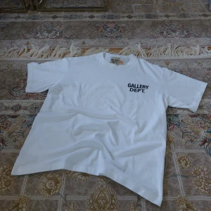 Vit t-shirt från Gallery Dept. - Vit t-shirt från Gallery Dept. med svart tryck på bröstet och stor text på ryggen där det står 'GALLERY DEPT. HOLLYWOOD, CA.'. Klassisk passform och rund hals. Tillverkad i mjuk bomull, perfekt för en clean och stilren look. Lapp inkluderad åt sidan, har du frågor meddela gärna 😊