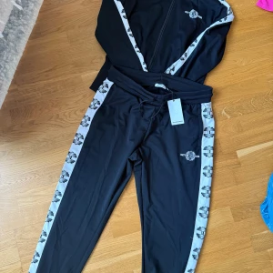 Svarta mjukisbyxor från Better Bodies - Svarta mjukisbyxor från Better Bodies med vita sidopaneler och svart-vitt mönster längs benen. Byxorna har snörning i midjan och broderad logga på ena benet. Perfekta för träning eller chill, tillverkade i mjukt syntetmaterial. Nypris 1500kr