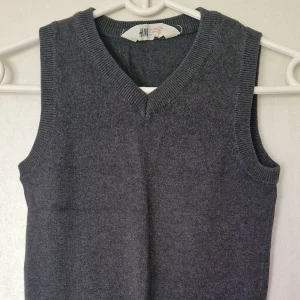 Mörkgrå finstickad väst från H&M - Mörkgrå stickad väst från H&M med v-ringning och ribbade kanter vid hals och ärmhål. Enkel och stilren modell som passar perfekt över en skjorta eller t-shirt för en snygg lager-på-lager-look.