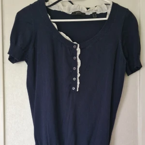 Mörkblå blus med volang från Esprit - Supersöt mörkblå blus från Esprit med korta ärmar, knappar framtill och vit volangdetalj längs halsringningen. Toppen är stickad i mjukt material och har en feminin touch med volangen och de små knapparna. Perfekt för dig som gillar en klassisk stil. Volangdetaljen behöver strykas lite på för att hamna rätt.
