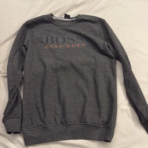 Hugo Boss - Snygg grå sweatshirt från Hugo Boss med stor BOSS-logga i mörkgrått och orange text framtill. Tröjan har rund halsringning, långa ärmar och ribbade muddar. Tillverkad i mjuk bomullsmix som är skön att ha på sig. Kan inye garantera äktheten på denna vara