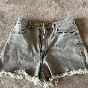 Grå jeansshorts med fransig kant - Säljer ett par grå jeansshorts med hög midja och rå, fransig kant nertill. Shortsen har klassisk femficksdesign, bälteshällor och dragkedja med knapp framtill. Perfekta för en chill sommarlook.