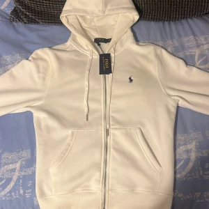Vit hoodie från Polo Ralph Lauren - Säljer en stilren vit hoodie från Polo Ralph Lauren med dragkedja och snörning i huvan. Tröjan har en broderad blå logga på bröstet, känguruficka och är tillverkad i mjuk bomull. Perfekt för en clean och avslappnad look.