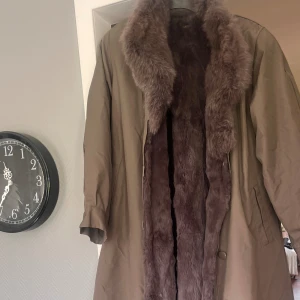 Lång pälsfodrad kappa Alexandra Päls - Säljer en lång och oversized kappa från Alexandra Päls. Yttertyget är grå khaki färgad  och insidan är fodrad med mjuk, grå päls. Kappan har bred krage, dolda knappar och rymliga ärmar. Perfekt för kalla dagar när du vill vara både varm och snygg.