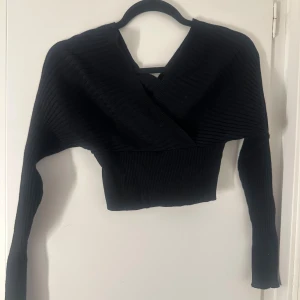 Svart croppad omlottstickad tröja - Svart stickad tröja med omlott-design och croppad passform. Tröjan har ribbad struktur och långa ärmar, perfekt för en trendig och stilren look. Djup v-ringning framtill ger en snygg detalj.