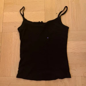 Svart linnetopp med rosett - En enkel och stilren svart linnetopp med smala axelband och en liten söt rosett framtill. Toppen har en rak passform och ärmlös design, perfekt att matcha med jeans eller kjol. Materialet känns mjukt och bekvämt mot huden.