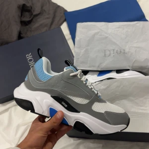 Chunky sneakers från Dior i grått och blått - Säljer ett par chunky sneakers från Dior med grått och vitt skinn, blå meshdetaljer och svarta samt blå inslag på sulan. Skorna har vit snörning och en modern, sportig vibe. Perfekta för dig som gillar exklusiva och trendiga sneakers.