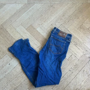 Blå jeans från Jacob Cohën, stl 34 - Säljer ett par klassiska blå jeans från Jacob Cohën i storlek 34. Jeansen har snygga kontrastsömmar, fem fickor och en diskret logga på myntfickan. Modellen är rak med normal passform och tillverkad i mjukt denimtyg för en skön känsla.