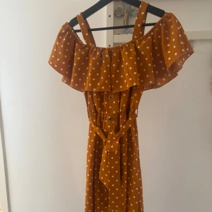 Prickig orange långklänning med volang - Superfin långklänning i orange med vita prickar, volang upptill och axelband. Klänningen har knappar framtill, markerad midja med knytband och är perfekt för sommarens alla tillfällen. Lätt och luftig känsla.