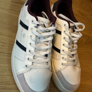 Vita sneakers från Gant med marinblå detaljer - Snygga vita sneakers från Gant med marinblå ränder på sidorna och vinröd insida. Skorna har snörning, rund tå och detaljer i mocka framtill. Klassisk och stilren design med Gant-logga på sidan och sula med mönster för bra grepp.