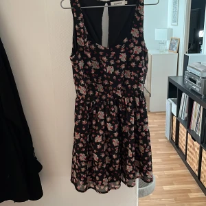 Blommig svart kortklänning Size s - Supersöt svart kortklänning från Size Needle med blommigt mönster i rosa och beige toner. Ärmlös modell med markerad midja och lätt utställd kjol. Perfekt för sommarens varma dagar och har en luftig känsla tack vare det tunna materialet.