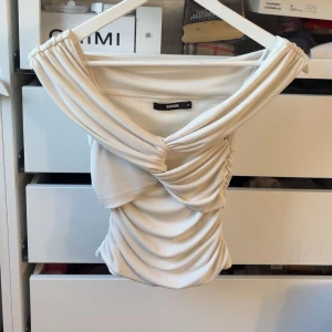 Offshoulder draperad topp från Bik Bok - Säljer en snygg offshoulder topp från Bik Bok i krämvit färg. Toppen har draperade detaljer framtill och baktill som ger en cool och trendig look. Gjord i mjukt stretchigt material och sitter tight mot kroppen. Perfekt för dig som vill sticka ut lite extra.