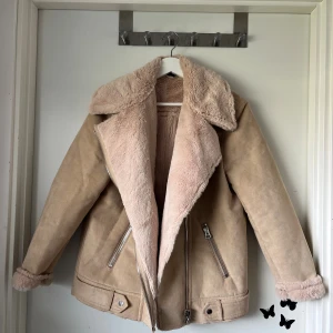 Beige teddyjacka med dragkedjor - Snygg teddyjacka i beige med fluffigt, ljusrosa foder och bred krage. Jackan har två silvriga dragkedjor framtill, detaljer vid midjan och uppvikta ärmslut med mjukt teddytyg. Perfekt för dig som gillar en chill och cozy stil under höst och vinter.