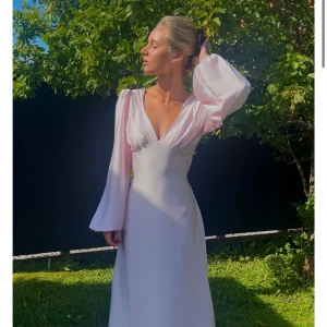 Ljusrosa långklänning med v-ringning - Superfin ljusrosa långklänning med djup v-ringning och vida långa ärmar. Klänningen har öppen rygg och knytband i midjan för en snygg siluett. Materialet är glansigt och faller mjukt, perfekt för dig som vill sticka ut med en romantisk look. Aldrig använt 