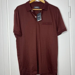 Vinröd polo t-shirt från Dressmann - Snygg vinröd polo t-shirt från Dressmann i mjuk bomull. Klassisk krage, korta ärmar med ribbade detaljer och en liten bröstficka med mesh.