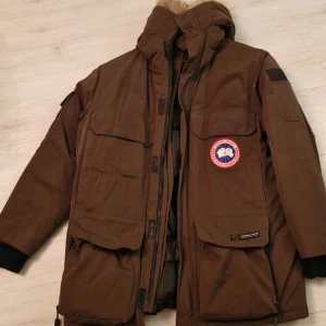 Brun Canada Goose dunjacka XS - Säljer en brun dunjacka från Canada Goose i storlek XS. Jackan har flera stora fickor, huva med pälsdetalj och klassisk logopatch på bröstet. Yttermaterialet är polyester och bomull, fodret är nylon och fyllningen är dun. Perfekt för kalla vinterdagar.