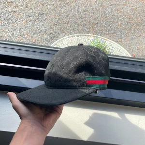 Svart Gucci keps med GG-mönster - Snygg svart keps från Gucci med diskret GG-monogram över hela kepsen. Klassisk böjd skärm och justerbar rem bak i skinn. På sidan finns en grön och röd rand som ger den typiska Gucci-looken. Materialet är bomull och skinn, perfekt för dig som gillar exklusiva accessoarer.