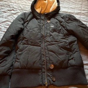 Svart pufferjacka Helly Hansen M - Svart pufferjacka från Helly Hansen med mysigt teddyfoder i huvan. Jackan har dragkedja, stora knappar och ribbade muddar. Klassisk HH-logga på bröstet och snygga detaljer på dragkedjan. Perfekt för kalla dagar när du vill hålla stilen.