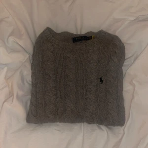 Grå kabelstickad tröja Polo Ralph Lauren - Kabelstickad grå tröja från Polo Ralph Lauren med klassisk rund halsringning och broderad logga på bröstet. Tröjan är i mjuk ull och har ribbade muddar vid ärmslut och nederkant. Perfekt för lager-på-lager under kyliga dagar.