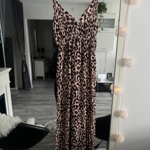 Leopardmönstrad långklänning New Yorker - Snygg långklänning från New Yorker med leopardmönster i beige och svart. Klänningen har v-ringning, smala axelband och resår i midjan för en bekväm passform. Perfekt för dig som vill sticka ut med ett djurmönstrat plagg.