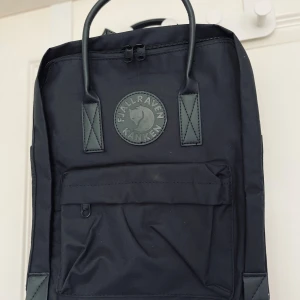Svart Fjällräven Kånken ryggsäck - Klassisk svart Fjällräven Kånken ryggsäck med ikonisk rund logotyp framtill. Rymligt huvudfack, dragkedja, framficka och justerbara axelremmar. Tillverkad i slitstark polyester och har praktiska handtag upptill. Perfekt för skola eller cityhäng. Fina läderdetaljer🖤 Helt i nyskick- aldrig använd! 