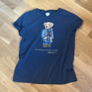 Blå Polo Bear t-shirt från Ralph Lauren - Mörkblå t-shirt från Polo Denim & Supply by Ralph Lauren med ikoniskt Polo Bear-tryck på bröstet. Klassisk passform med rund hals och korta ärmar. Tillverkad i mjuk bomull, perfekt för en chill och stilren look.
