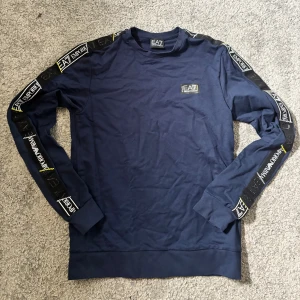 Blå långärmad tröja från EA7 - Snygg marinblå långärmad tröja från EA7 Emporio Armani med logga på bröstet och tryck längs ärmarna. Tröjan har rund halsringning och är gjord i mjuk bomull, perfekt för en stilren och sportig look.