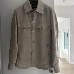 Beige linnéovershirt från Oscar Jacobson - Stilren beige overshirt från Oscar Jacobson i linne. Jackan har klassisk krage, knäppning framtill med fyra knappar, två bröstfickor med lock och knappar samt raka ärmslut med knappdetalj. Perfekt för lager-på-lager och en clean look.
