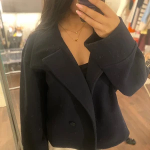 Mörkblå ullkappa med bred krage - Snygg mörkblå kappa i ull med bred krage och knäppning framtill. Jackan har en rak, lite boxig passform och långa ärmar. Perfekt för dig som gillar stilrena och klassiska plagg med en modern twist.