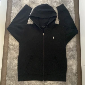 Svart hoodie från Polo Ralph Lauren L - Svart hoodie från Polo Ralph Lauren i storlek L Klassisk design med dragkedja framtill, huva och fickor. Liten vit broderad logga på bröstet. Perfekt för en chill och stilren look. Det står att det är Storlek Xl men eftersom att jag har använt den och har haft den i ett tag så passar det även för dem som har Storlek L så rekommenderar den starkt för de som har Storlek L