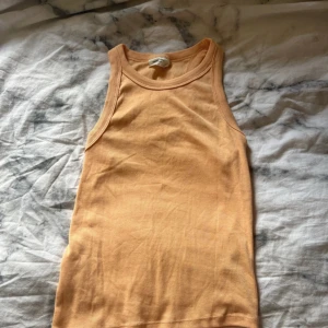 Persikofärgad ribbad linnetopp Gina Tricot - Säljer en persikofärgad ribbad linnetopp från Gina Tricot, basically basics-kollektionen. Toppen är ärmlös med klassisk rund halsringning och har en tight passform. Perfekt att styla med jeans eller kjol för en enkel och fräsch look.