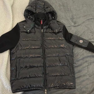 Svart dunväst med huva från Moncler - Snygg svart dunväst från Moncler med glansig finish, avtagbar huva och dragkedja framtill. Västen har ribbstickade svarta ärmar och en ikonisk Moncler-logga på ärmfickan. Perfekt för lager-på-lager och streetstyle.