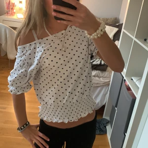 Prickig tröja/blus - Säljer en söt vit blus med svarta prickar. Toppen har korta puffärmar med resår, croppad passform och smockad volangkant nertill. Den har en fyrkantig ringning och kan bäras off-shoulder för en trendig look. Perfekt för dig som gillar en lekfull och stilren vibe. Den är från Gina men lappen är inte kvar . Man kan även ha den på lite olika sätt som ni ser på bilderna !Ingen aning om storlek men passar XXS- XS-S. Hör av er vid funderingar !💖💖💖
