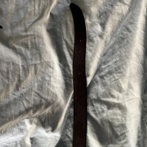 Brunt mockabälte från Onewear Sweden - Snyggt brunt bälte i mocka från Onewear Sweden med stilren silverfärgad metallspänne. Bältet har en klassisk rak form och kommer med originalpåse. Perfekt för att lyfta din outfit med en sofistikerad touch.