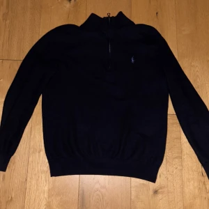 Mörkblå stickad tröja Polo Ralph Lauren - Mörkblå stickad tröja från Polo Ralph Lauren med halv dragkedja och broderad logga på bröstet. Tröjan har lång ärm, ribbade muddar och klassisk passform. Perfekt för lager-på-lager och snygg till jeans eller chinos.