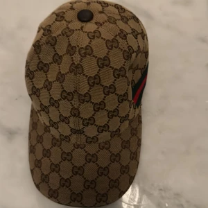 Gucci keps med GG-mönster och ränder - Exklusiv keps från Gucci i beige med klassiskt GG-monogram över hela ytan. Sidan har den ikoniska gröna och röda randen. Justerbar rem i skinn baktill och insida i bomull. Perfekt för dig som vill sticka ut med lyxig streetstyle. Pris kan diskuteras 