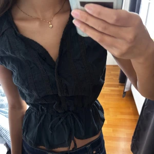 Svart blus med knyt från Yessica - Svart blus från Yessica med v-ringning, knappar framtill och fina spetsdetaljer. Modellen har korta ärmar och knyt i midjan för en snygg siluett. Tillverkad i mjuk bomull, perfekt till jeans eller kjol.