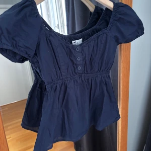 Mörkblå blus från Hollister - Supersöt mörkblå blus från Hollister med kort puffärm, v-ringning och knappar framtill. Modellen har resår vid halsringning och midja för en snygg passform. Perfekt till jeans eller kjol för en chill look.